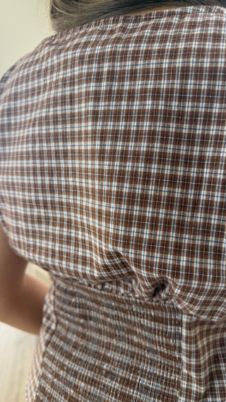 Close-up of a brown and white plaid garment worn by a person, showing the back of the shirt or dress with a small button closure near the lower back.