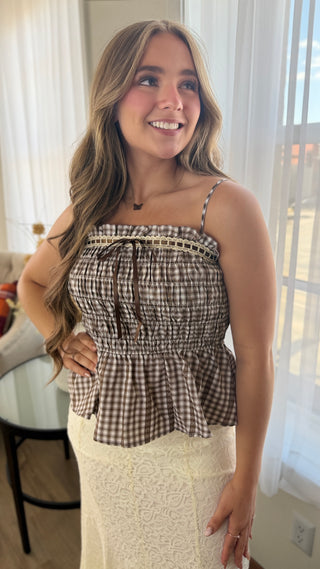 A woman wearing a brown and beige gingham smocked peplum top with thin spaghetti straps and a cream lace midi skirt, standing in a brightly lit room with white curtains.