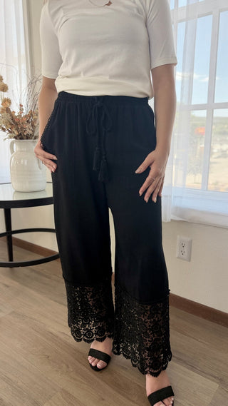 A person wearing loose black wide-leg pants with a drawstring waist and black lace trim at the hems, paired with a white short-sleeve top and black sandals in a bright indoor room.