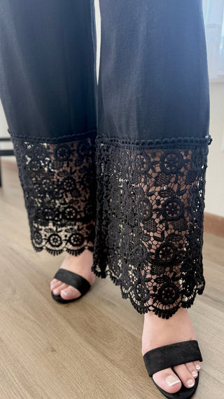 Black wide-leg pants with a detailed floral lace hem, shown on a model wearing black slide sandals on a light wood floor.