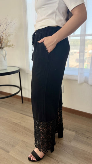 A person wearing a long black maxi skirt with a lace hem and side pockets, paired with a white top and black slide sandals, standing in a bright room with wood floors and a window.