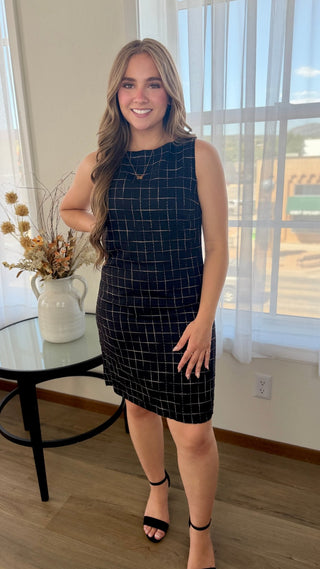 A woman modeling a sleeveless black dress with a white windowpane grid pattern, standing in a brightly lit room with a window and a small side table.