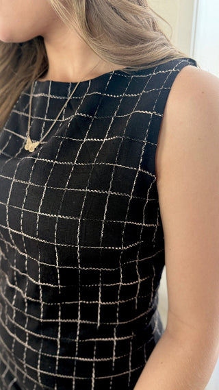 Close-up of a woman wearing a sleeveless black top with a white windowpane grid pattern and a delicate gold necklace.