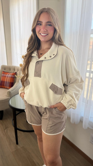 A woman modeling a cream and taupe two-piece lounge set consisting of a long-sleeve quarter-placket pullover and matching shorts, standing in a brightly lit room with sheer curtains.
