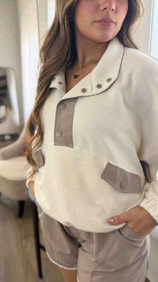 A woman wearing a cream and taupe color-blocked pullover top with snap-button placket and matching shorts in a bright indoor setting.