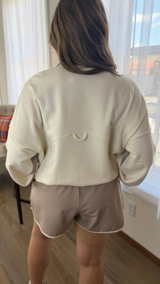 A woman viewed from behind wearing a cream-colored cropped sweatshirt and taupe shorts in a brightly lit room with a window and wooden floor.