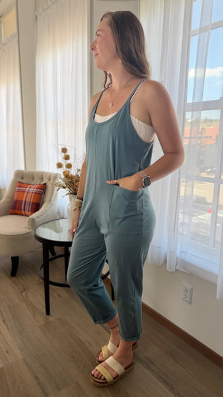 A woman wearing a dusty blue sleeveless jumpsuit over a white tank top, standing in a bright room with large windows and sheer curtains.