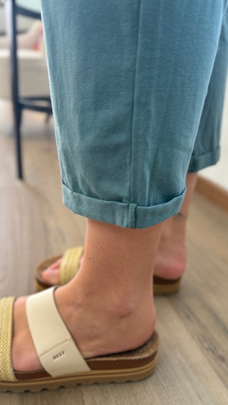 A close-up of a person's feet wearing light blue cuffed pants and cream-colored slide sandals with brown soles, standing on a light wood floor.