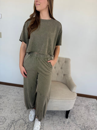 Woman wearing an olive green outfit standing in a room with a chair and white wall.
