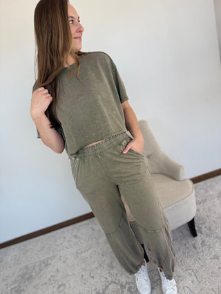 Woman wearing an olive green outfit standing in a room with a chair and light-colored walls.