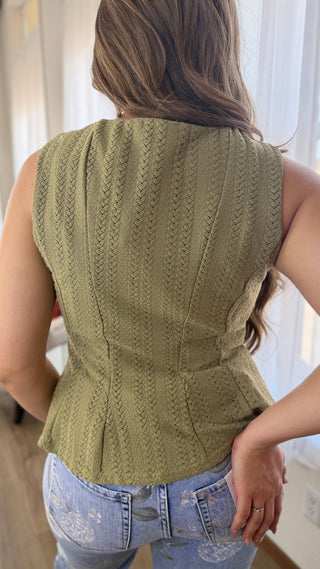 A woman viewed from behind wearing a sleeveless olive green textured knit top with a gathered peplum waist, paired with light blue jeans.