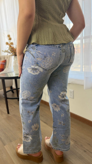 A person shown from the back wearing light-wash blue jeans with a floral embroidered pattern, paired with a textured olive green top and brown platform sandals in a home interior setting.