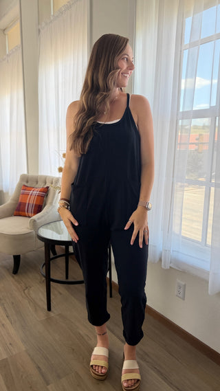 A woman modeling a black sleeveless jumpsuit with a white undershirt, standing in a brightly lit room with large windows and sheer curtains.