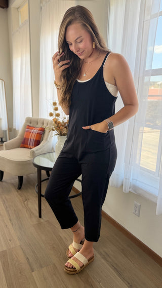 A woman modeling a black sleeveless jumpsuit with a white underlayer in a bright room with wood floors, white curtains, and a beige armchair.