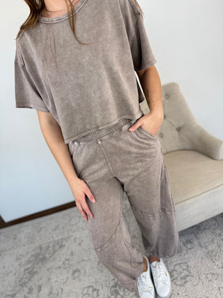Person wearing a brown oversized t-shirt and pants in a room with a beige chair.