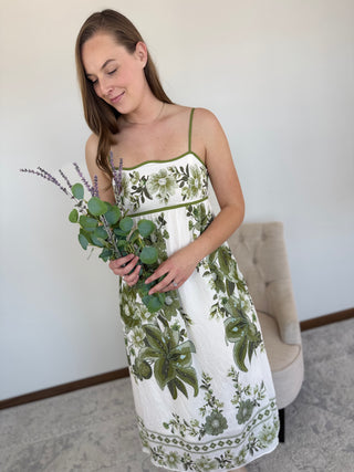Woman in a white dress with green floral patterns holding a bouquet of flowers indoors.