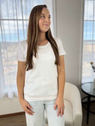 Woman wearing a white t-shirt and light pants standing in a room with large windows.