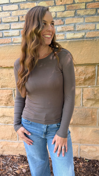 A woman with long wavy brown hair stands outdoors against a tan brick wall, wearing a fitted long-sleeve taupe-brown top and light blue jeans.