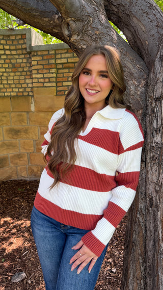 A woman wearing a red and white striped ribbed knit polo sweater and blue jeans, standing outdoors next to a large tree trunk.