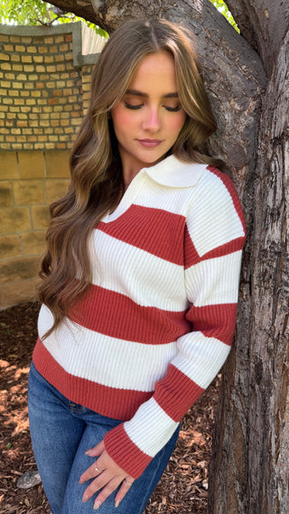 A woman with long wavy brown hair leans against a tree trunk outdoors, wearing a ribbed knit sweater with wide horizontal red and off-white stripes, paired with blue jeans.