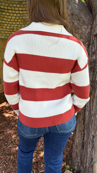 A person viewed from behind wearing a ribbed knit sweater with wide horizontal red and off-white stripes, paired with blue jeans, standing outdoors next to a tree trunk.