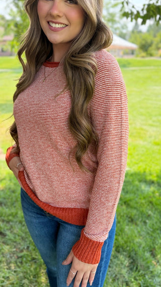 A woman wearing a textured knit crewneck sweater in a heathered coral pink and rust orange color, paired with blue jeans, standing outdoors on a grassy lawn.
