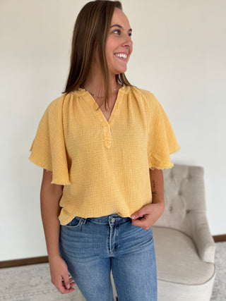 Woman wearing a yellow blouse and blue jeans standing in a room with a neutral background.