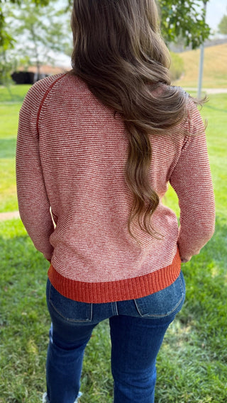 A woman viewed from behind wearing a textured, heathered coral-pink knit sweater with a contrasting rust-orange ribbed hem, paired with blue jeans, standing outdoors on a grassy lawn.