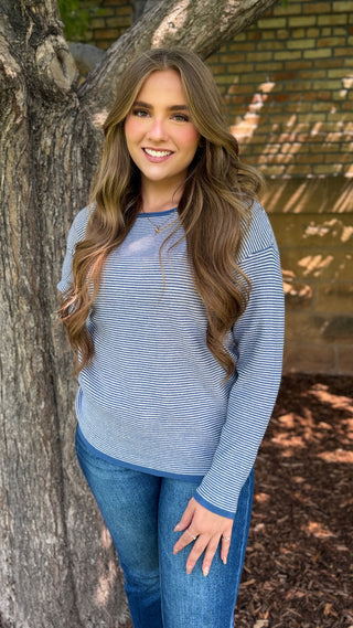 A woman with long wavy brown hair stands outdoors next to a tree trunk, wearing a light blue and white horizontally striped long-sleeve top with blue jeans.