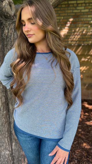 A woman wearing a light blue and white striped long-sleeve top with solid blue trim, paired with blue jeans, standing outdoors next to a tree.