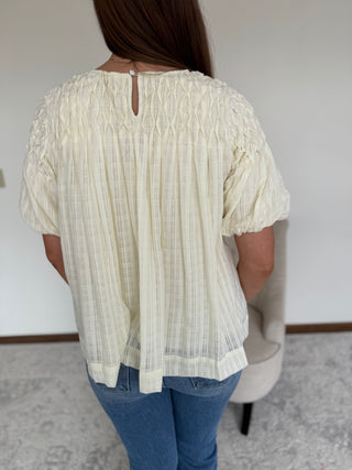Person wearing a light-colored blouse with short sleeves and blue jeans in a room.