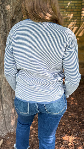 A person viewed from behind wearing a light blue and white striped long-sleeve top with blue jeans, standing outdoors next to a tree trunk.