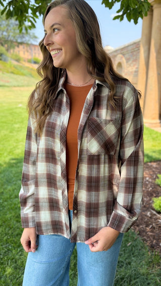 A woman wearing a brown, beige, and white plaid button-up flannel shirt over an orange-brown top and blue jeans, standing outdoors in a grassy area.