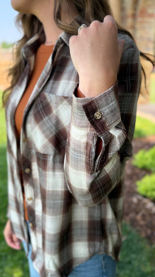 A woman wearing a brown, beige, and white plaid button-down shirt, shown from the side in an outdoor setting.
