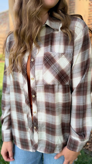 A woman wearing a brown, beige, and white plaid button-up flannel shirt with blue jeans, photographed outdoors.