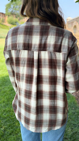 A person viewed from behind wearing a brown and cream plaid flannel shirt and blue jeans, standing outdoors on a grassy lawn.