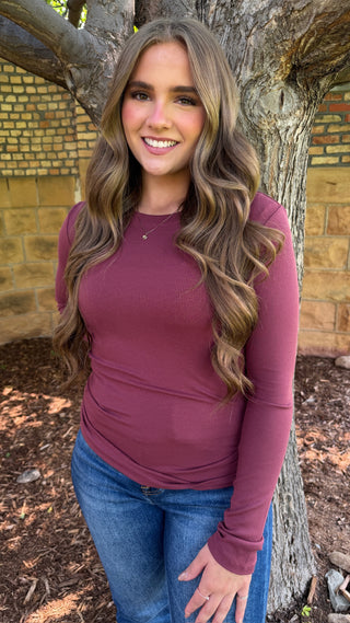 A woman with long wavy brown hair stands outdoors against a tree trunk, wearing a fitted long-sleeve crewneck top in a muted plum-burgundy color and blue jeans.