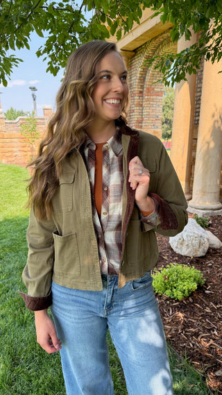 A woman wearing an olive green utility jacket with a brown corduroy collar, layered over a brown and beige plaid shirt and blue jeans, standing outdoors in front of a brick building with columns.