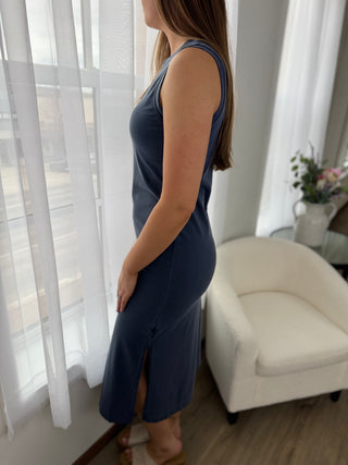 Woman wearing a blue dress standing in a room with white curtains and a white chair.