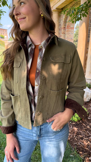 A woman wearing an olive green utility jacket with brown faux-fur trim on the cuffs, layered over a brown and white plaid flannel shirt and an orange top, paired with blue jeans in an outdoor setting.
