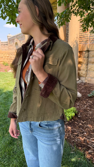 A woman wearing an olive green utility jacket with a dark brown corduroy collar and matching brown corduroy cuffs, layered over a plaid shirt and blue jeans, standing outdoors in a garden setting.
