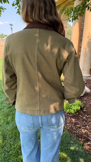 A person viewed from behind wearing an olive green canvas jacket with a brown corduroy collar, paired with light blue jeans, standing outdoors on a grassy lawn.