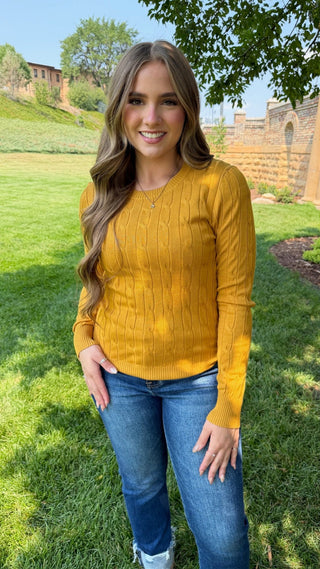 A woman standing outdoors on a grassy lawn wearing a mustard yellow long-sleeve cable-knit sweater and blue jeans.