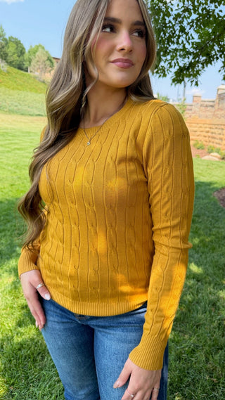 A woman wearing a mustard yellow cable-knit sweater and blue jeans, standing outdoors on a grassy lawn with trees and hills in the background.