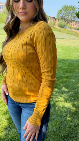 A woman wearing a mustard yellow cable-knit sweater and blue jeans, standing outdoors on a grassy lawn.