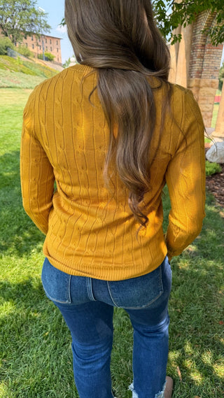 A woman viewed from behind wearing a mustard yellow cable-knit sweater and blue jeans, standing outdoors on a grassy lawn.