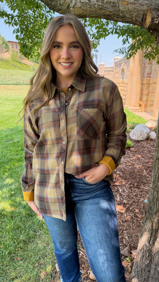 A woman stands outdoors under a tree wearing a long-sleeve plaid button-up shirt in brown, beige, and muted red tones, paired with blue jeans.