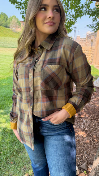 A woman wearing a long-sleeve plaid button-up shirt in olive green, brown, and tan, paired with blue jeans, standing outdoors on a grassy lawn.
