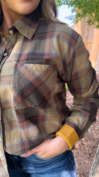 A close-up of a person wearing a long-sleeve plaid flannel shirt in muted brown, beige, and blue tones, paired with blue jeans. The shirt features a chest pocket and a ribbed yellow cuff.