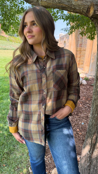 A woman wearing a brown and beige plaid flannel button-up shirt with yellow ribbed cuffs, standing outdoors under a tree next to a brick building.
